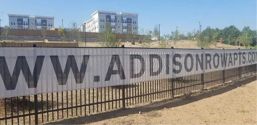 Large printed advertising banner attached to a construction-site fence.